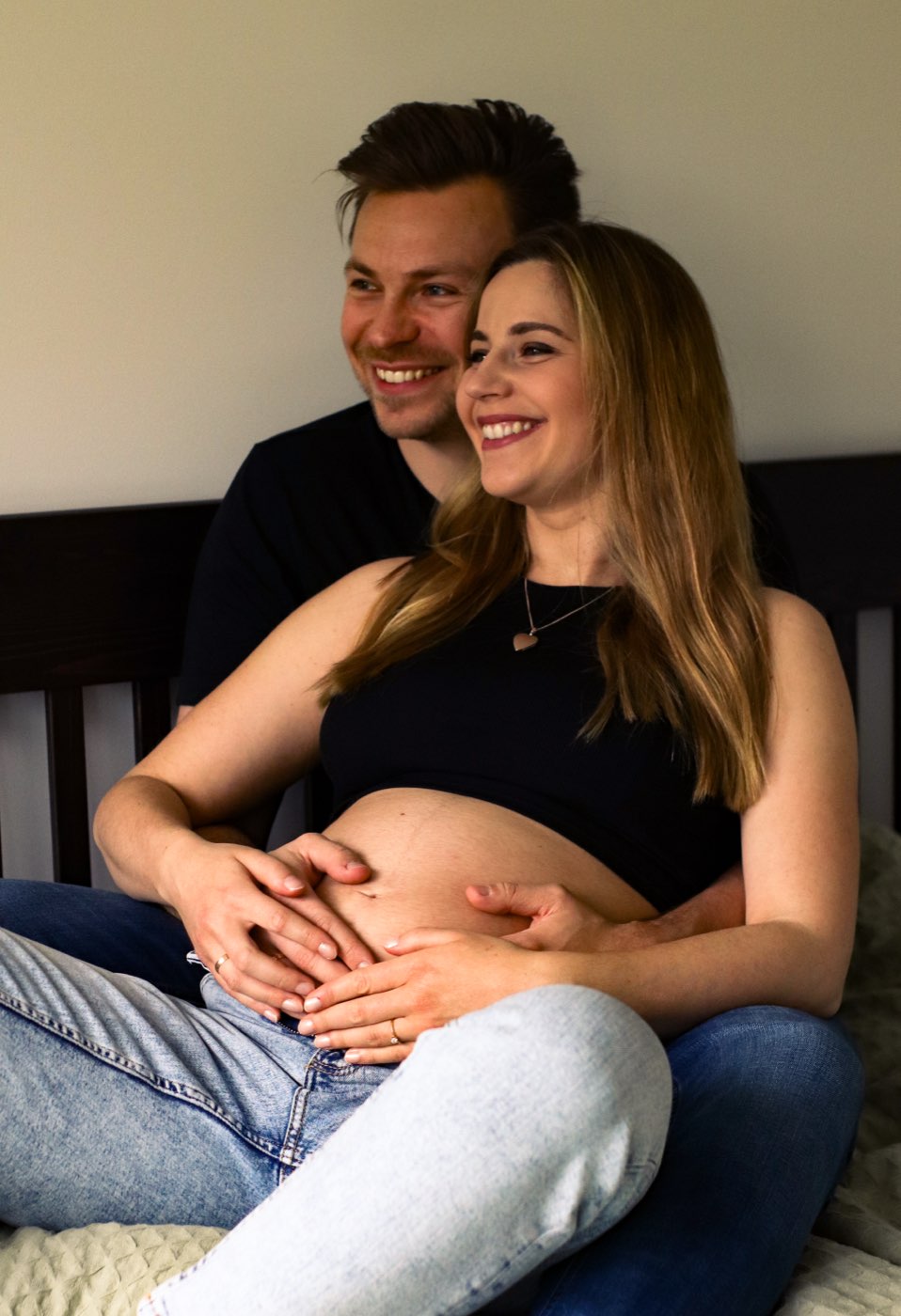 Ein werdendes Elternpaar sitzt lachend auf dem Bett, die Hände auf dem Babybauch