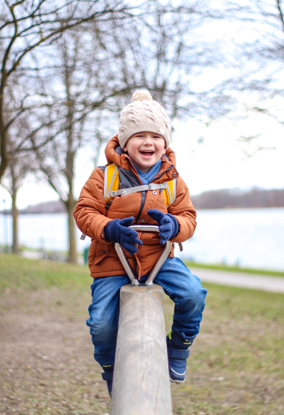 Ein Bub mit Haube und oranger Jacke lacht auf einer Wippe