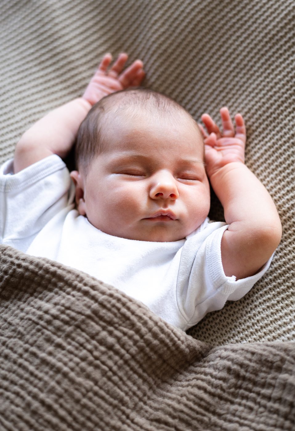 Nahaufnahme eines schlafenden Babys mit erhobenen H&auml;ndchen, halb zugedeckt
